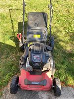 Front view of Briggs & Stratton lawn mower showing red deck and black engine housing with label visible. Grass collection bag is attached at the rear.