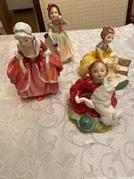 Four porcelain figurines on tablecloth: woman in pink/red dress (Goody Two Shoes), woman in yellow dress with basket, woman playing with dog, and woman in green and white dress with headscarf.