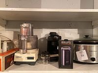 Photo showing Cuisinart food processor, Hamilton Beach crockpot, boxed Krups coffee grinder, and a black coffee machine placed on a white shelf.