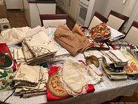 Photo showing a large assortment of table linens on a dining table including napkins, tablecloths, embroidered pieces, and dish towels all stacked and folded.
