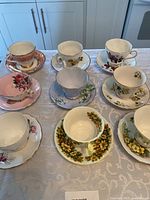 Full display of seven teacups and matching saucers in various floral patterns and colors on a white tablecloth