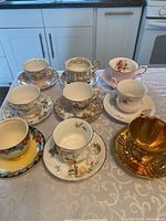 Photo showing seven vintage teacups and saucers arranged on a table, displaying various floral and decorative patterns with gold trims.