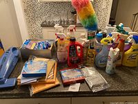Full view of cleaning supplies on the counter including spray bottles, gloves, wipes, cloths, sponges, and a rainbow feather duster.