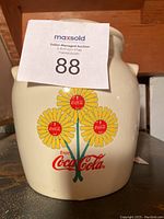 Front view of ceramic cookie jar showing floral Coca-Cola design, cream color, and lid knob partially visible.