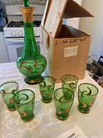 Green glass decanter and six sherry glasses with swirl and gold designs arranged on a white tablecloth with the original cardboard box in the background.