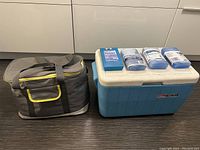 Blue Coleman cooler with white lid and red logo, four assorted freezer packs on top, and gray soft cooler bag with yellow trim beside cooler on dark floor.