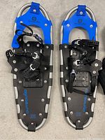 Top view of pair of snow shoes showing blue top, black foot platform, adjustable black bindings, and metal cleats underfoot. Brand 'Outbound' visible on foot platform with size '25'.