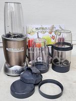 Photo of the NutriBullet Magic Bullet motor base, three blending containers including one tall, one medium with handle, and one smaller, plus multiple lids and recipe booklet on white background.