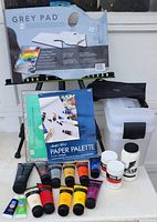 Photo showing acrylic paint tubes of various colors, two paper pads (Grey Pad and Paper Palette), a black metal folding easel, and two storage boxes.