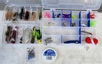 Photo showing two clear plastic tackle boxes with compartments. One box contains assorted handmade fishing flies and various fishing lures, some in feather and synthetic material. Other box contains soft plastic lures, small fishing hooks, swivels, and two red spherical bobbers. A spool of Scientific Anglers fly line backing and small fishing hooks in a box are in front of the tackle cases.