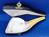Full view of the Meerschaum pipe nestled inside fitted black case with white satin lining, showing the pipe's bowl carved with face and attached yellow stem.