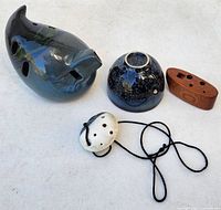 Four ocarinas displayed on a white surface showing different shapes and materials: dark blue pig-shaped ceramic, black speckled bowl-shaped ceramic, white ceramic with pendant cord, and small wooden ocarina.