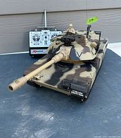 Front view of M1A2 remote control tank showing long barrel, camouflage paint, mounted figure, and Speed remote control behind the tank.