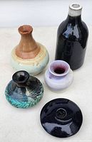 Five pottery bud vases arranged on a surface showing diverse colors and shapes including black and cream, teal and orange, teal and black, lavender and cream, and blue vases.