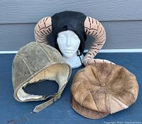 Lot of three hats, showing all the included items on a dark flat surface, hats include black leather and horned Tim the Enchanter style, worn shearling leather aviator cap, and brown patchwork flat cap.