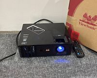 Front view of projector powered on next to its box and remote