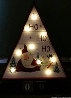 Front view of triangular advent calendar with LED lights turned on showing 'HO HO HO' and festive painted figures (Santa Claus and snowman) with lit bulbs.