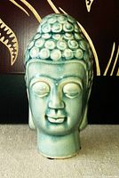 Close-up photo of olive-colored ceramic Buddha head figurine with smooth surface and detailed features.