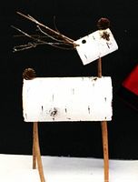 White birch log reindeer decorated with pinecones for nose and antlers, standing on four wooden legs, photographed against a black backdrop on a white surface.