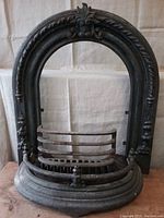 Full assembled view of largest decorative arched fireplace surround with attached fire grate and fender guard on wooden floor against cloth background.