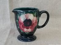 Side view of Moorcroft creamer showing floral decoration and handle against a neutral fabric background.