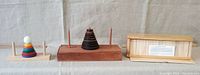 Three wooden puzzles displayed side by side: the multi-colored Tower of Hanoi puzzle with rings and white ball, the natural wood circular ring Hanoi Tower, and the wooden block puzzle in a wooden box