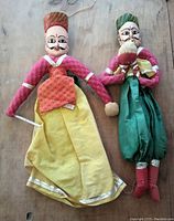 Two Rajasthani puppets lying side by side on wood surface. One in yellow and red, the other in green and red holding a bird.