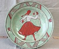 Front view of a circular ceramic plate with pale green background and a hand-painted dancing woman in a red skirt holding a flower, geometric border and floral highlights.