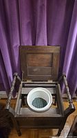 Front view of antique wooden commode with top lid open revealing white ceramic chamber pot inside, set against a purple curtain background.
