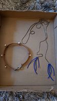 Photo showing both the leather choker with pearls and the silver chain necklace with blue beadwork tassels placed flat in a cardboard box.