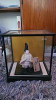 Full view of Asian figurine inside clear glass display case on a purple rug, with wooden background behind the case.