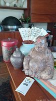 View of the plaster Buddha statue, metal pot, red hand-painted container, and blue flower pot with fan