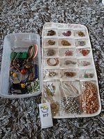 Photo showing the hanging jewelry organizer pouch with multiple pairs of brooches, earrings, and two necklaces below, alongside a plastic container with additional bracelets and jewelry pieces inside.