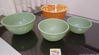 Four vintage Fire King nesting bowls displayed on a black surface: three green and one orange bowl.
