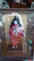 Full view of the vintage Japanese doll displayed inside a wooden framed glass case. The doll wears a red and orange traditional kimono with floral detail and holds a silver fan.
