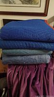 Stack of four folded professional moving blankets in various shades of blue, with one blanket having an orange border visible.