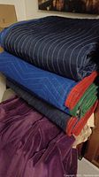Stack of four moving blankets showing folded edges in blue with white stitching and color edge bindings (red, green, orange) plus purple soft blanket underneath.