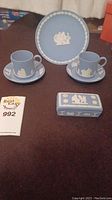 Photo showing two blue jasperware cups and saucers with white relief designs, a matching 6.5 inch plate with classical motif, and a rectangular lidded box with white decorative relief.