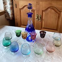 Full display of blue lead crystal decanter with stopper, four colorful cut crystal stemmed glasses, and ten small colored glasses in various hues arranged on white tablecloth.