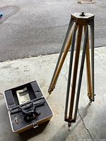 Metal tripod standing on ground with the Craftsman transit-level and its case placed beside it