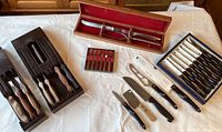 Overview showing two black plastic storage organizers, eight-piece Cutco steak knife set, three loose knives, Gerber carving set in wooden box and small colored-handled tool block