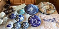 Photo of multiple ceramic and porcelain East Asian inspired dishes and bowls in shades of blue, white, and brown, laid out on a table with embroidered dragon textiles visible.