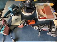 Lot showing Makita finishing sander, buffer/polisher, Skil orbit sander, Bosch random disc sander, and sandpaper sheets in a tray on dark surface.