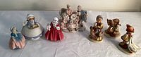 Seven porcelain figurines arranged on a table showing full lot including Baroque couple, Royal Doulton, Coventry Ware, and Hummel figurines.