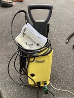 Front view of yellow Karcher pressure washer with power cord and hose wrapped on unit, showing handle and attached manual.