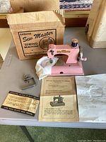 Full shot of pink sewing machine with clamp, original box behind it, instruction sheet and handwritten note laid out on table