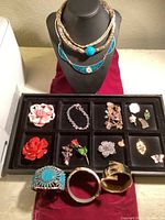 Photo showing a black jewelry display bust with layered gold-tone and blue enamel necklace, two wide cuff bracelets in front, and a black tray with assorted pins and small jewelry pieces.