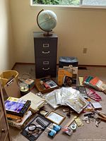 Photo showing black filing cabinet with vintage globe on top, assorted office supplies including photo paper, paper clips, calculator and scissors around base.