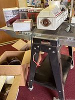 Drum sander mounted on black stand with caster wheels, with sandpaper boxes and loose rolls visible on table surface.