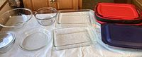 Overview of all glass bakeware and storage containers on white tablecloth including bowls, pie pan, rectangular baking dishes, and lids in red, black, and blue.